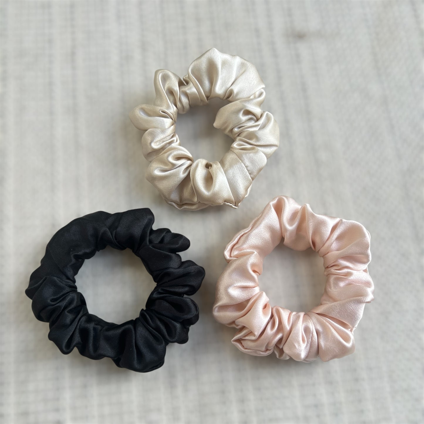 Three silk scrunchies in black, champagne, and pink on a textured surface