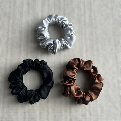 Three silk scrunchies in silver, black, and brown on a textured surface.