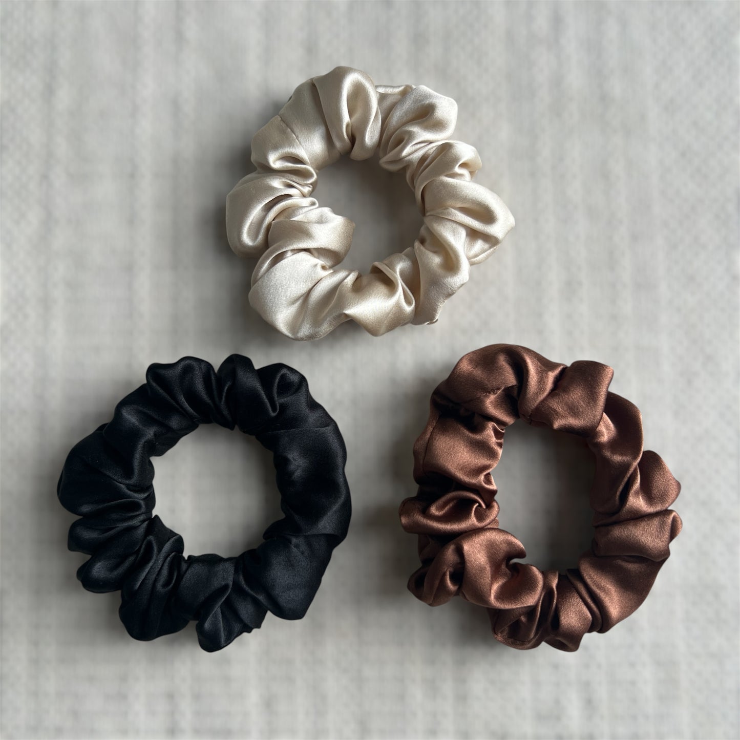 Three silk scrunchies in champagne, black and brown on a textured background