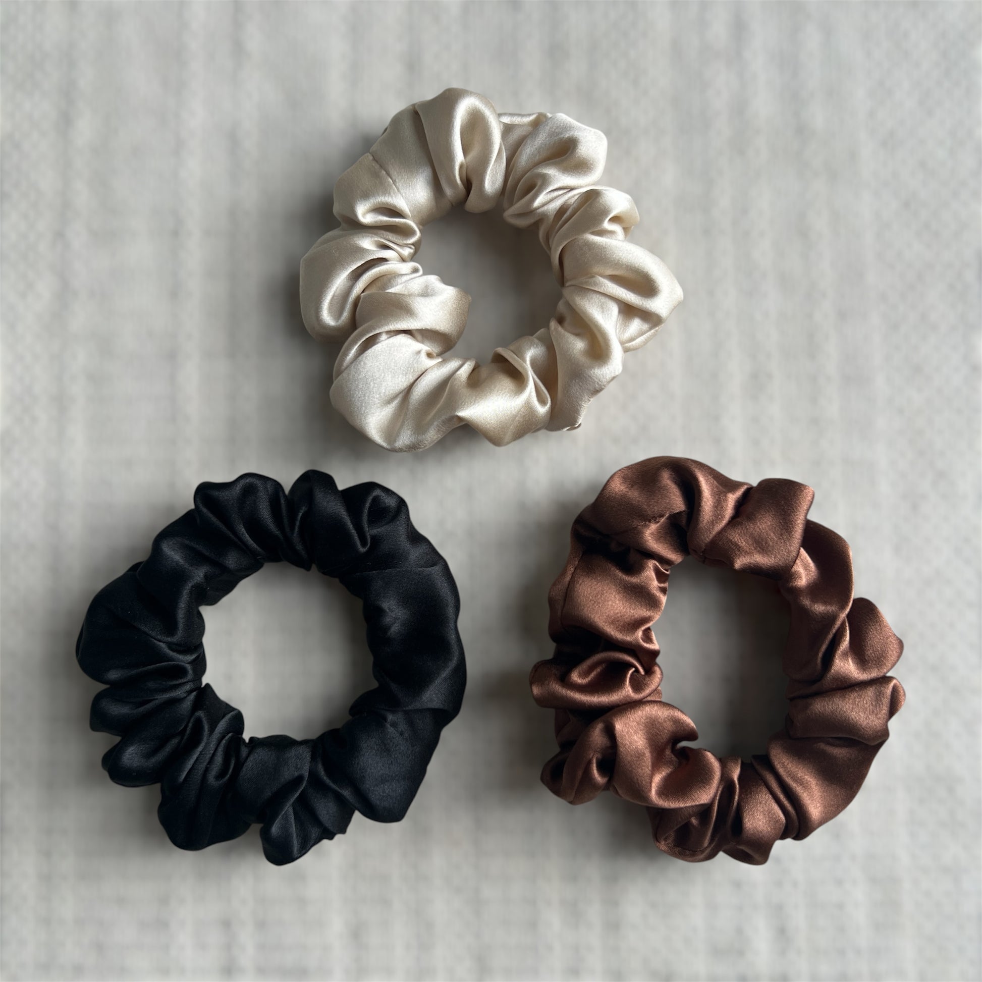 Three silk scrunchies in champagne, black and brown on a textured background