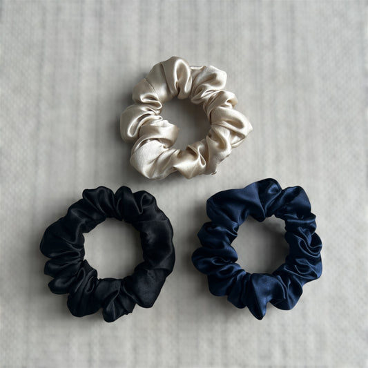 Three silk scrunchies in black, champagne, and navy on a textured surface.