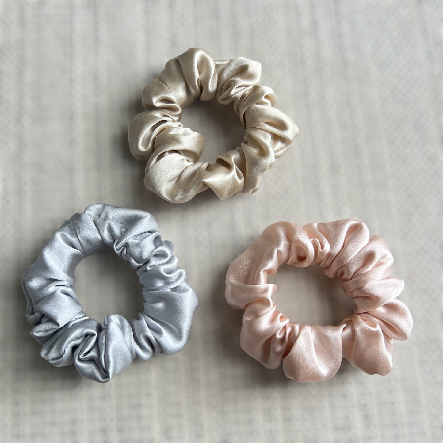 Three silk scrunchies in champagne, silver and pink on a textured surface