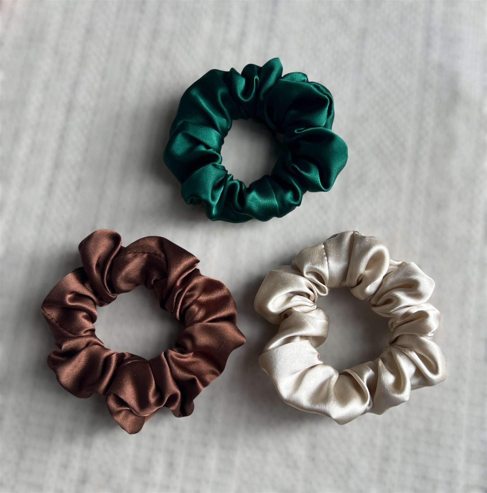 Three satin scrunchies in green, brown, and beige on a textured surface.