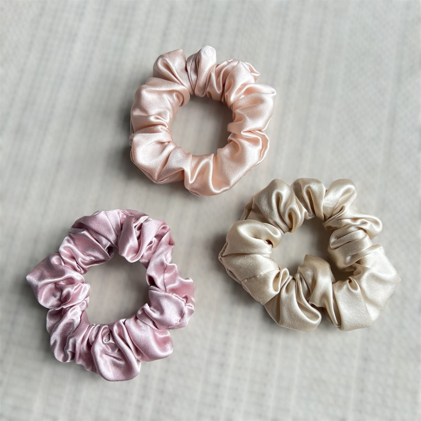 Three silk scrunchies in pink, rose gold, and champagne on a textured surface.